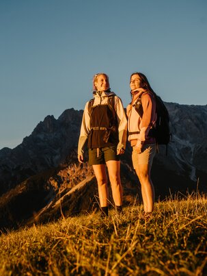 Two female hikers stand on a grassy slope in evening light in Dienten with a rocky mountain massif near Gabühelbahn. | © Hochkönig Bergbahnen