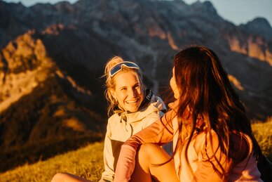 Zwei Wanderinnen sitzen im Abendlicht in Dienten vor einem felsigen Bergmassiv nahe der Gabühelbahn. | © Hochkönig Bergbahnen