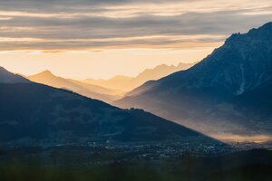 View over Dienten across a valley with mountain silhouettes, sun rays and a cloud-filled sky in morning light. | © Hochkönig Bergbahnen