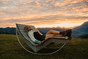 A woman lies on a wooden lounge bench on a grassy slope in Dienten with mountain views at sunset. | © Hochkönig Bergbahnen