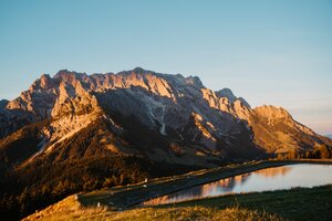 Sunlight illuminates a rocky mountain massif above a reservoir in Dienten beneath a clear evening sky. | © Hochkönig Bergbahnen