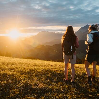 Two female hikers stand on a grassy slope in Dienten looking at mountains near Gabühelbahn at sunset. | © Hochkönig Bergbahnen