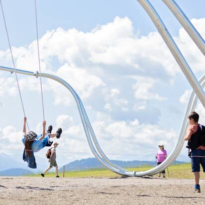 Kids swinging on oversized swings at Hochkönig swing park with Alpine backdrop on a sunny day | © Manu Lochner