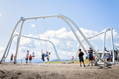 Kinder schaukeln auf überdimensionalen Schaukeln im Schaukelpark Hochkönig vor Alpenkulisse bei Sonnenschein | © Manu Lochner