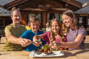 Four people sit at a wooden table outside an alpine hut and share a dish with salad in the sun. | © Hochkönig Bergbahnen, Roland Haschka