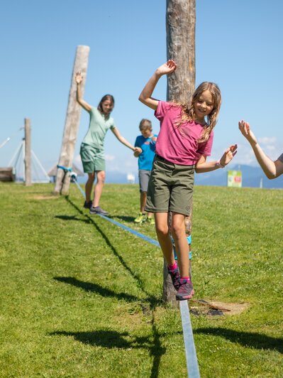 Ein Mädchen balanciert auf einer Slackline in Mühlbach, während ein Mann hilft und zwei Kinder folgen. | © Hochkönig Bergbahnen, Roland Haschka