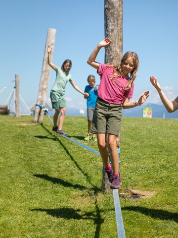 Ein Mädchen balanciert auf einer Slackline in Mühlbach, während ein Mann hilft und zwei Kinder folgen. | © Hochkönig Bergbahnen, Roland Haschka