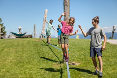 A girl balances on a slackline in Mühlbach while a man helps and two children follow behind. | © Hochkönig Bergbahnen, Roland Haschka