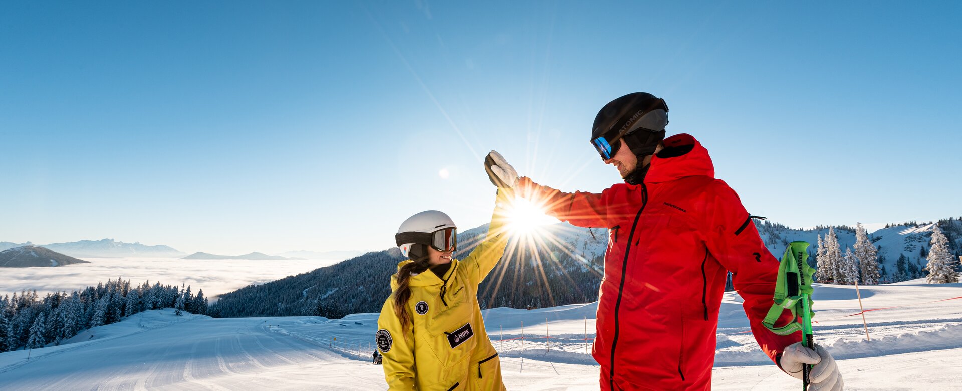 Zwei Skifahrer in bunter Kleidung geben sich ein High-Five auf sonniger, frisch präparierter Skipiste