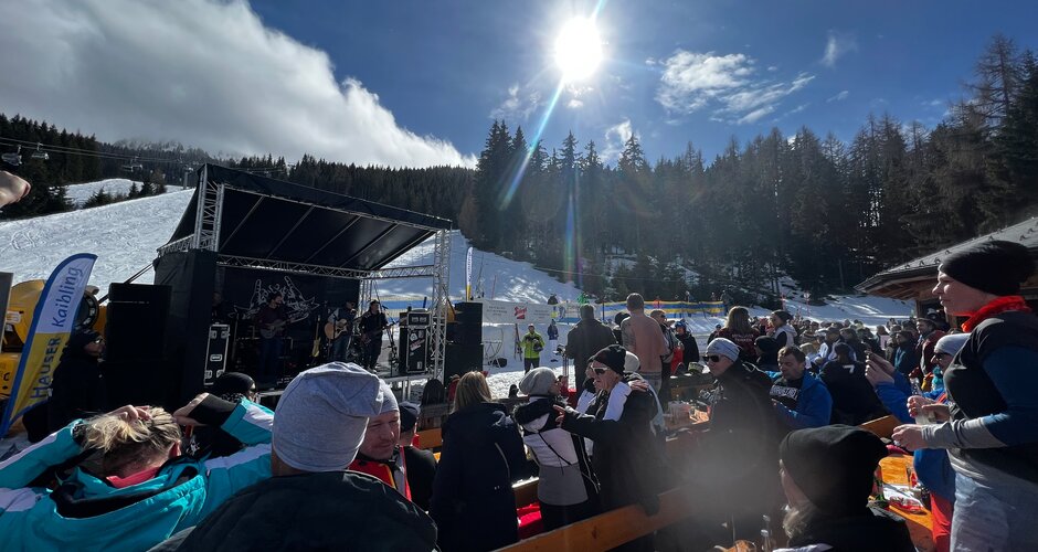 Viele Personen in Skiausrüstung stehen oder sitzen auf der Skihütten-Terrasse und blicken auf eine kleine Bühne auf der Musiker spielen | © Hauser Kaibling