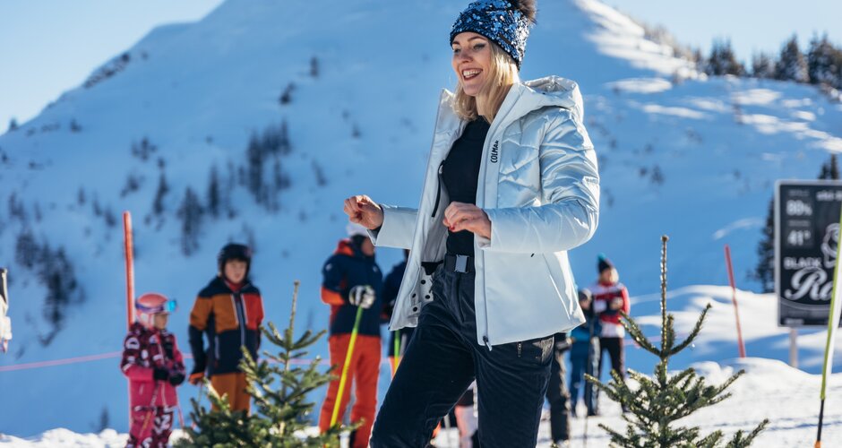 Wintermode mit Bergkulisse auf der Reiteralm bei Sonnenschein | © Gerald Grünwald Frau in heller Winterjacke posiert lächelnd im Schnee, im Hintergrund Berge und Skifahrer | © Gerald Grünwald