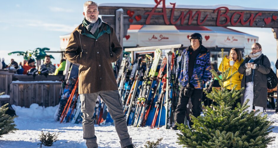 Wintermode vor der Almbar auf der Reiteralm bei Sonnenschein | © Gerald Grünwald Ein Mann posiert in Wintermode vor der Almbar, im Hintergrund Skier, Gäste und Schneelandschaft | © Gerald Grünwald