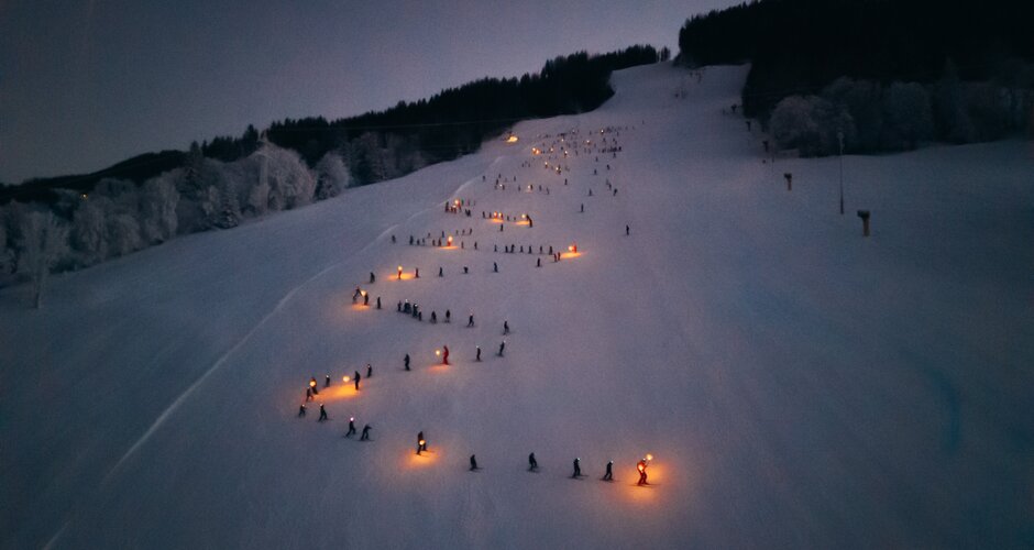 Kinder fahren mit Fackeln bei Dunkelheit die Reiteralm hinab, orange Lichter im Schnee sichtbar | © Gerald Grünwald