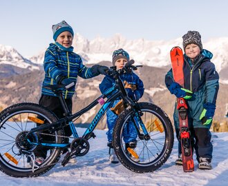 Drei Kinder stehen im Schnee, eines mit Fahrrad, eines mit Ski, Bergpanorama im Hintergrund | © Gerald Grünwald