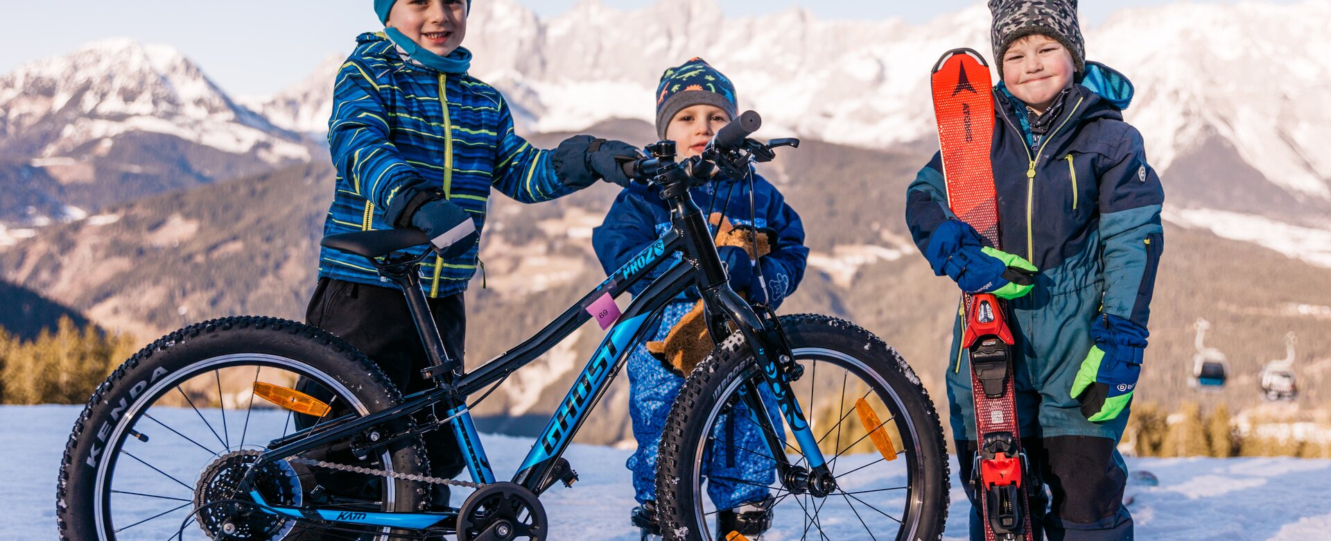 Drei Kinder stehen im Schnee, eines mit Fahrrad, eines mit Ski, Bergpanorama im Hintergrund | © Gerald Grünwald