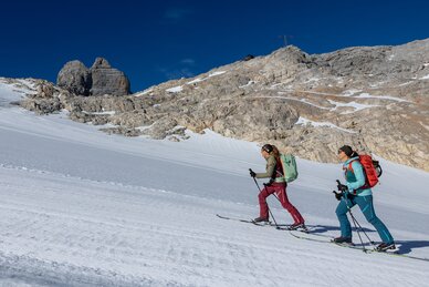 Zwei Frauen steigen mit Tourenski einen verschneiten Hang unter Felswänden hinauf | © Harald Steiner