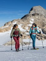 Zwei Frauen gehen mit Skitouren-Ausrüstung vor felsigem Gipfel bei Sonnenschein | © H. Steiner
