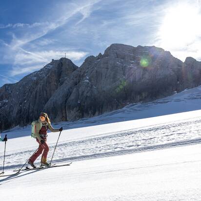 Zwei Frauen steigen mit Tourenski bei Sonne vor steiler Felswand im Schnee bergauf | © Harald Steiner
