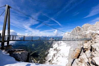 Personen überqueren Hängebrücke am verschneiten Dachstein, mit weitem Blick ins grüne Ennstal unter blauem Himmel. | © Harald Steiner