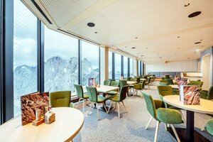 Helles Restaurant mit großen Panoramafenstern, grünen Polsterstühlen und Holztischen mit Blick auf die umliegenden Berge. | © Christoph Huber