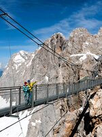 Zwei Personen überqueren die Hängebrücke am Dachstein mit Blick auf schneebedeckte Gipfel und schroffe Felslandschaft bei Sonne. | © Harald Steiner