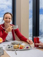 Lachende Frau in roter Jacke trinkt Kaffee, Mann mit rotem Getränk, Alpenpanorama durch große Fenster, Dessert auf Tisch. | © Harald Steiner
