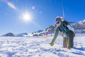 Woman kneeling in snow with probe and avalanche transceiver, sun and mountains in the background. | © René Eduard Perhab