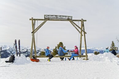 Skitourengeher sitzen auf großer Holzschaukel mit Ausblick auf verschneite Bergwelt