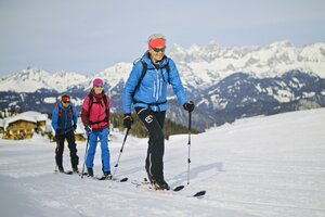 Drei Skitourengeher steigen lächelnd auf der Piste mit Blick auf verschneite Berge auf