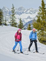 Zwei Skitourengeherinnen gehen bei Sonne auf breiter Spur, verschneite Berge im Hintergrund