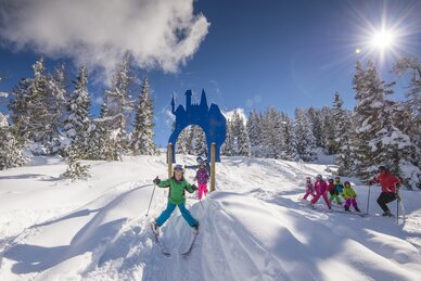 Kinder fahren Ski durch ein Tor in Schlossform, umgeben von verschneiten Bäumen. | © Lorenz Masser