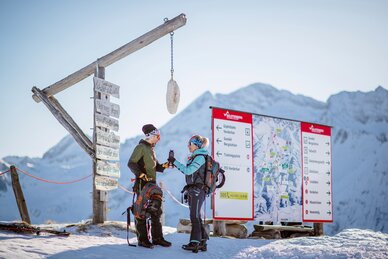 Zwei Wintersportler stehen an einer Wetterstation mit Holzschild in den Bergen und studieren eine große Pistenkarte. | © Christoph Huber
