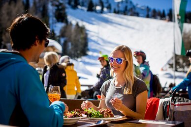 Frau mit Sonnenbrille genießt Essen auf der Skihütte bei Sonnenschein, Skifahrer im Hintergrund. | © Tom Lamm