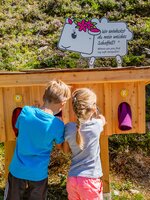Zwei Kinder tasten in einer hölzernen Fühlstation nach Stoffen, Schild mit Schaf stellt Rätsel auf dem Themenweg. | © Hauser Kaibling