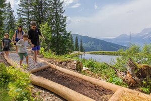 Familie mit Kindern barfuß auf einem Naturpfad mit Kies und Erde am Hauser Kaibling, im Hintergrund ein Bergsee und Alpenpanorama. | © René Eduard Perhab