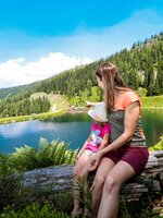 Frau mit Kind sitzt auf Baumstamm am klaren Bergsee, beide schauen auf das Wasser, umgeben von Wald und Wanderweg. | © Stefan Pajman