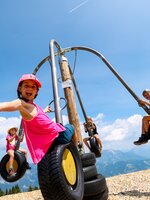Kinder schwingen auf einem Karussell aus Reifen vor Bergkulisse, lachen und strecken die Arme fröhlich in die Kamera. | © Stefan Pajman