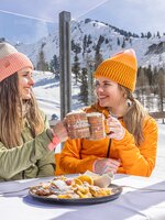 Zwei Frauen mit Mützen stoßen mit Getränken an, Teller mit Kaiserschmarrn im Vordergrund. | © Bernhard Moser