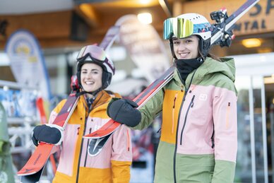 Zwei Skifahrerinnen in bunter Skikleidung mit Skiern vor Skiverleih, bereit für den Skitag. | © Bernhard Moser