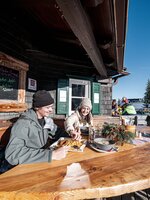 Zwei Personen essen draußen an einer Skihütte mit Holzbank und Schneehintergrund. | © Hauser Kaibling