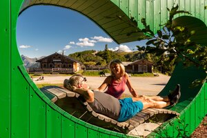Wanderpaar entspannt auf Herzbankerl aus Holz mit Blick auf Almhütten und sonnige Berglandschaft. | © Christine Höflehner