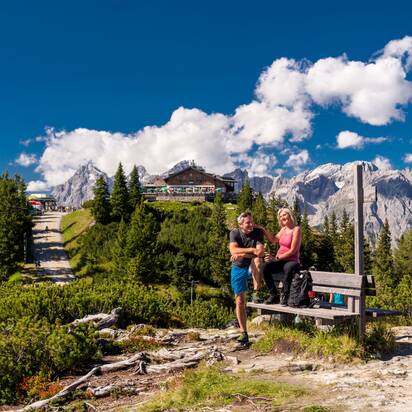Wanderpaar macht Rast auf Bank mit Blick auf Hochwurzenhütte und markante Felsgipfel bei Sonnenschein. | © Christine Höflehner