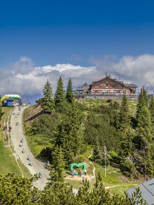 GoKart-Fahrer und Wanderer am Gipfel der Hochwurzen mit Almhütte, Herzplatz und Panorama unter blauem Himmel. | © Planai/Kovacsics