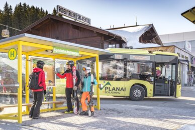 Skifahrer mit Ausrüstung an der Skibusstation Hochwurzen, Skibus im Hintergrund. | © David Stocker