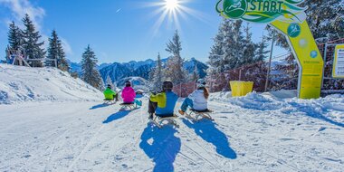 Four sledders start on the Hochwurzen toboggan run on a sunny winter day with mountain backdrop. | © René Eduard Perhab