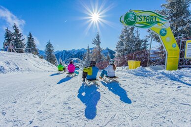Four sledders start on the Hochwurzen toboggan run on a sunny winter day with mountain backdrop. | © René Eduard Perhab
