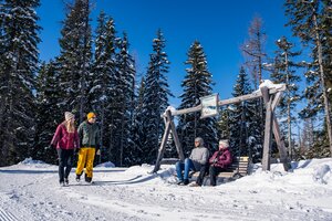Winterwanderer und sitzende Gäste auf einer Holzbank vor verschneiten Bäumen. | © Gerald Oberreiter