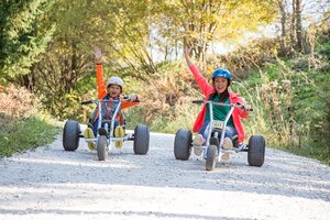 Frau und Kind fahren nebeneinander mit MountainKarts auf Schotterweg und heben lachend die Arme. | © Elisabeth Wolkenstein