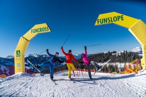 Three skiers in colorful outfits happily posing at the start of the Funslope and Funcross track in a sunny ski resort. | © Christine Höflehner