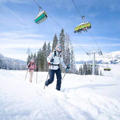 Zwei Skitourengeherinnen steigen durch den Schnee, während über ihnen ein Sessellift mit Skifahrern fährt. | © Josh Absenger
